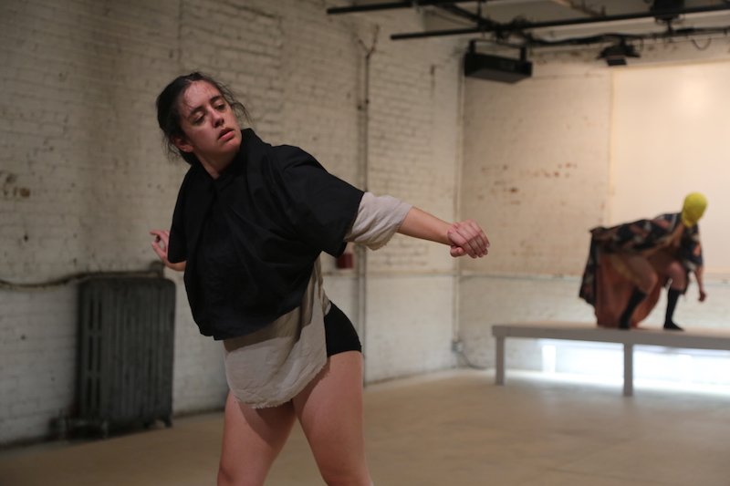A woman in black and beige tunic peers in front of her. A masked dancer crouches on a raised platform in the background
